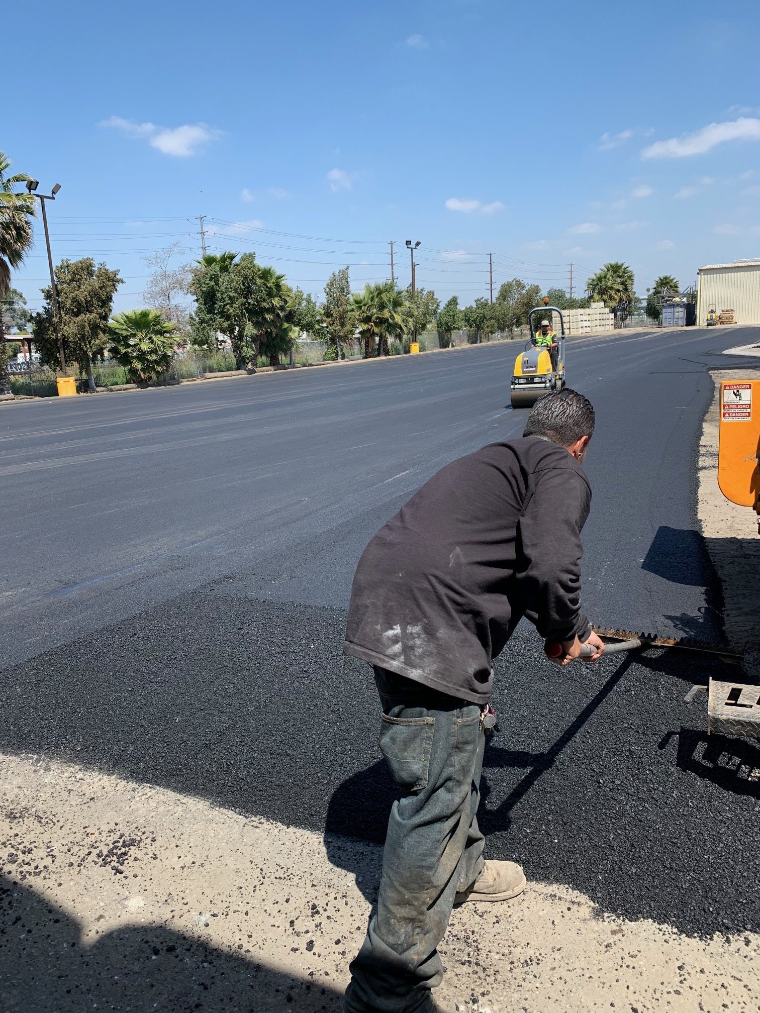 Professional worker raking hot mix asphalt while an asphalt roller compacts a newly paved road by Black Diamond Asphalt SoCal.